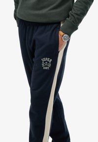 Marineblauwe sweatpants met beige zijstrips, voorzien van een logo met "SUPER DRY" en oprichtingsdatum. Elastische tailleband, ontspannen pasvorm, katoenen materiaal.