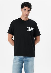 T-shirt de algodão preto com decote redondo, mangas curtas e um logótipo branco com letras em negrito no lado esquerdo do peito. Usado com jeans de lavagem clara.