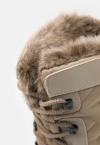 Beige vinterstøvle med faux fur krave, tekstureret stof, sort hardware, snørebånd design og forstærket syning langs kanterne.