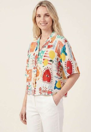 Donna sorridente con capelli biondi che indossa una camicia colorata a maniche corte con motivi astratti rossi, gialli e blu e pantaloni bianchi.