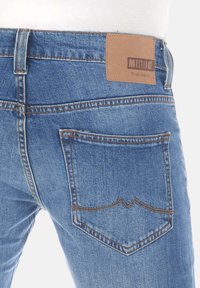 Los jeans de mezclilla azul claro presentan un parche de cuero marrón claro con la marca "MUSTANG", bolsillos estándar y detalles de costura sutiles en la parte posterior.