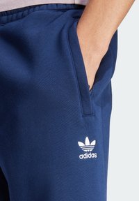 Navy modré tepláky vyrobené z bavlněné směsi, s boční kapsou a bílým logem Adidas se třemi pruhy nad ním.