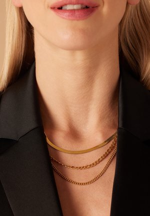 Trois colliers superposés : une chaîne en or plate, une chaîne en or de taille moyenne et une fine chaîne en or, reposant contre un haut à col sombre.