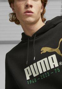 Sudadera negra con bolsillo canguro, con un gran logo blanco de "PUMA", acentos dorados y números verdes: 1948-2023-75. Material suave.
