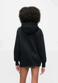 Person med krøllete hår iført svart hettegenser og grønne shorts, vist bakfra mot en ensfarget hvit bakgrunn.