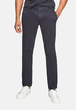 Mann trägt dunkelblaue, gerade geschnittene Hose mit Seiten taschen und weiße Sneaker mit niedrigem Schaft, steht mit einer Hand in der Tasche.