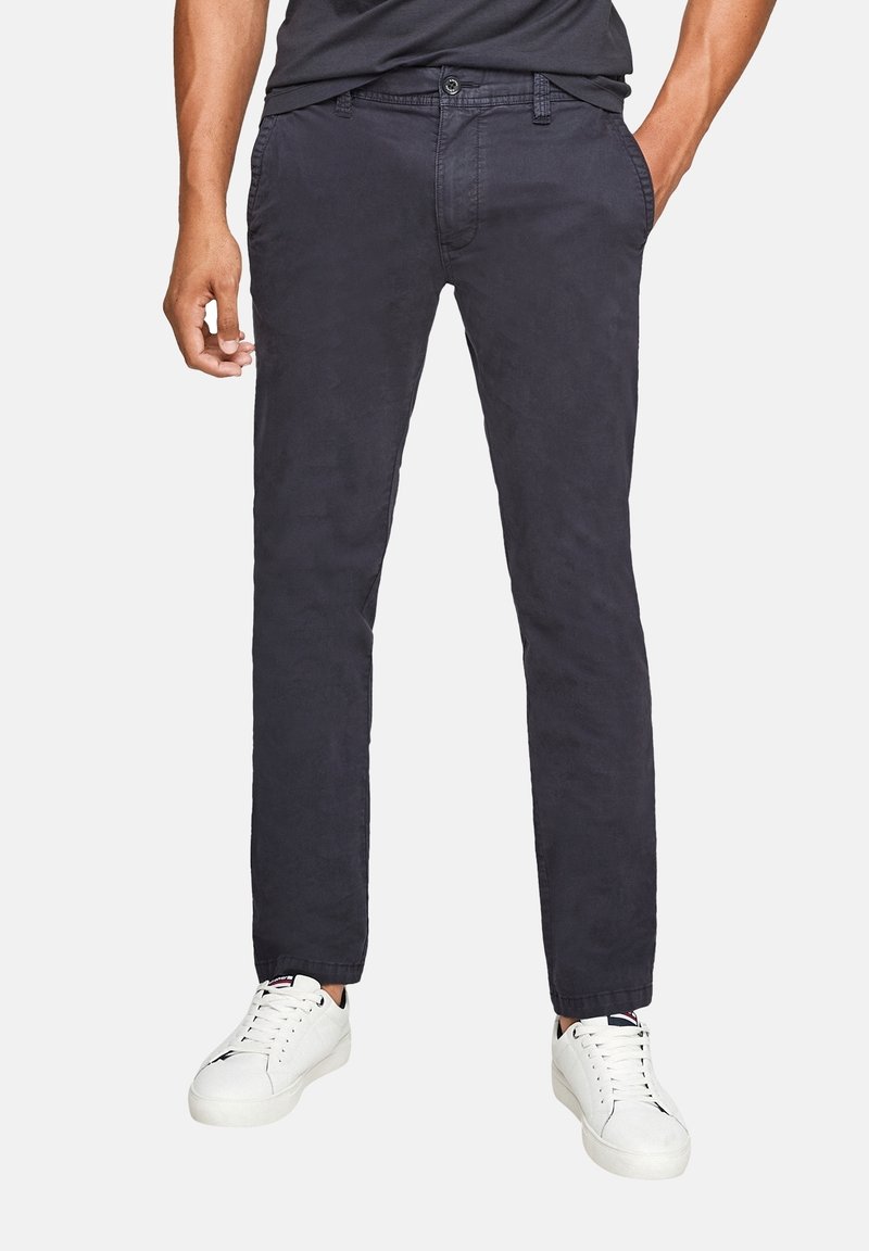 Homme portant un pantalon droit bleu marine foncé avec poches latérales et des baskets basses blanches, debout avec une main dans la poche.