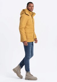 Gul isolerad jacka med huva, två framfickor och dragkedjestängning, ihop med blå jeans och grå snörkängor.