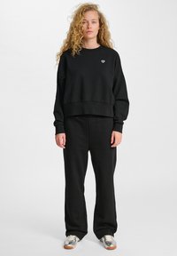 Schwarzes Crewneck-Sweatshirt mit einem verkürzten Design, das ein kleines weißes Logo zeigt. Kombiniert mit locker sitzenden schwarzen Jogginghosen und silbernen Sneakers.