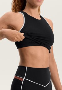 Čierny športový top s bielym lemovaním, priliehavým dizajnom a priedušnou textúrou. Osoba si upravuje tkaninu pri páse.