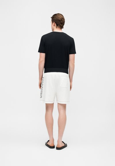 Άνδρας που φαίνεται από πίσω, φορώντας μαύρο πουκάμισο με κοντό μανίκι, λευκά σορτς με κείμενο στο πλάι και μαύρα σανδάλια, στέκεται σε λευκό φόντο.