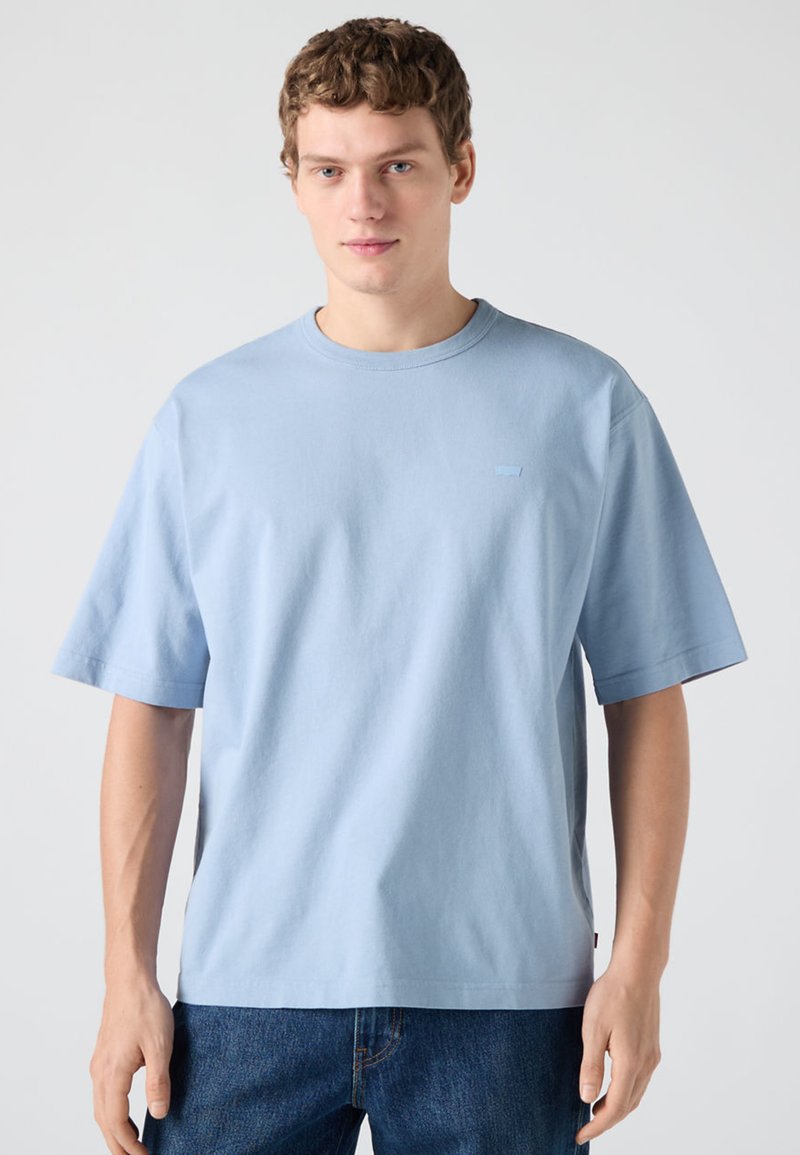 Νεαρός άνδρας με κοντά σγουρά μαλλιά, φορώντας μπλε ανοιχτό oversized κοντομάνικη μπλούζα και σκούρα μπλε τζιν, στέκεται μπροστά σε απλό φόντο.