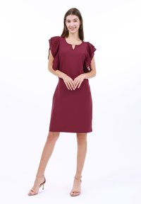 Burgundy klänning med fladdrande ärmar, rund halsringning och nyckelhålsdetalj. Slät textur med en knälång silhuett. Bärs med nudefärgade klackar.