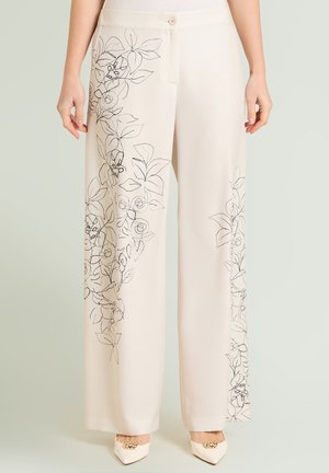 Pantaloni a gamba larga color crema con disegni floreali neri a linee lungo le gambe, vita con bottoni, indossati con tacchi bianchi a punta con dettagli dorati.