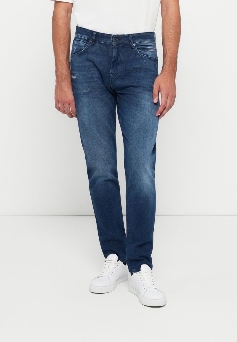 Jeans din denim albastru închis cu o croială dreaptă, având detalii subtile de decolorare și efect de whiskering, asortați cu adidași albi.