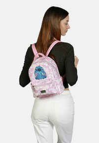 Mochila de tela rosa con patrón floral y gráfico de personaje azul. Cuenta con correas ajustables, cierre con cremallera y bolsillo frontal con herrajes.
