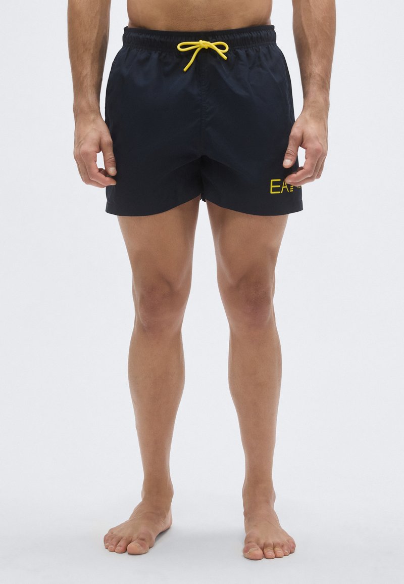 Tengerészkék úszónadrág sárga zsinórral, rugalmas derékrésszel és kis sárga logóval az oldalon. Könnyű, sima anyagból készült, térdig érő fazon.