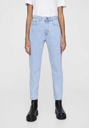 Personne portant un jean droit taille haute bleu clair, un top blanc à manches courtes et des bottines noires à plateforme épaisse, debout devant un fond uni.