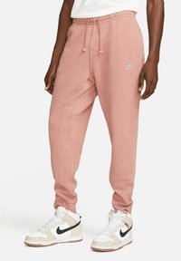 Nike joggers in light pink cotton blend with elastic waistband, drawstring, side pockets, and logo detail near hip. Pairs with white and beige sneakers.