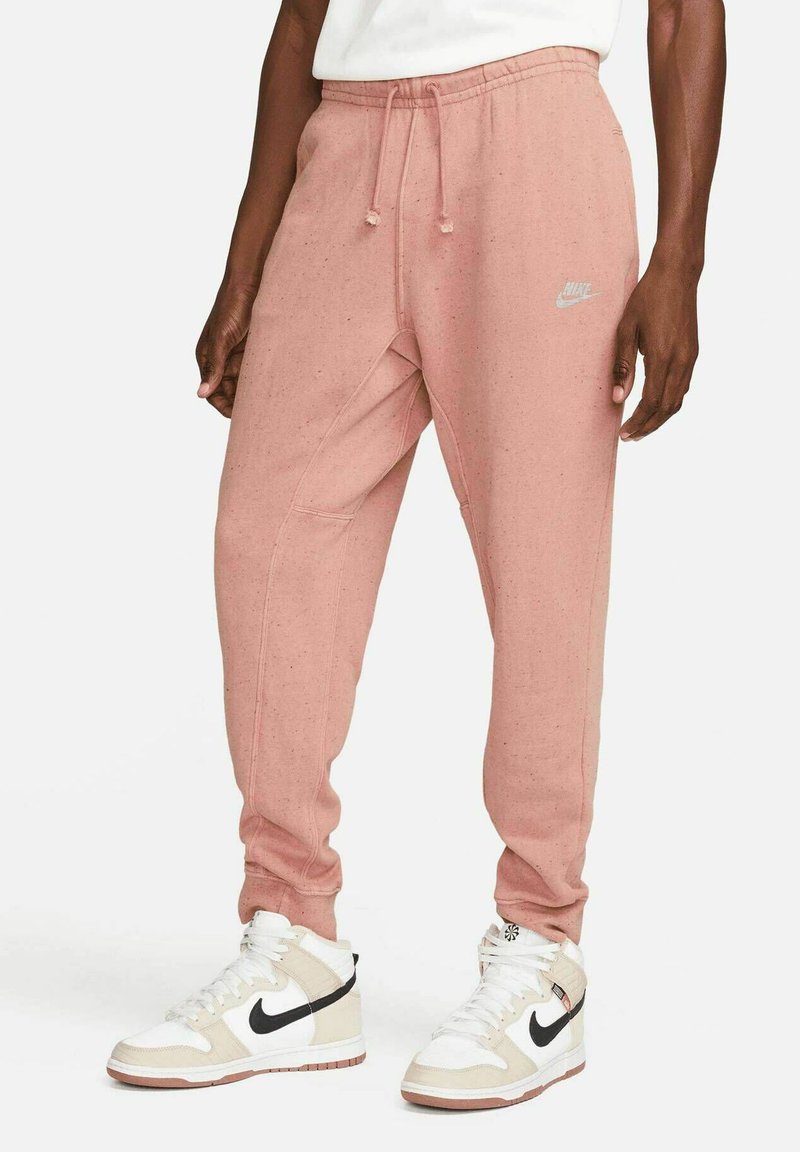 Nike joggers in light pink cotton blend with elastic waistband, drawstring, side pockets, and logo detail near hip. Pairs with white and beige sneakers.