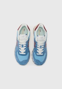 Light blue sneakers with suede and mesh upper, white laces, red accents on the sides, and a black rubber sole. Model: New Balance 574.