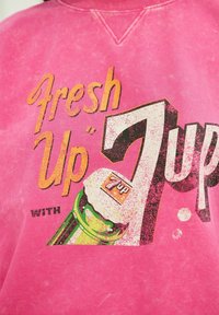 Lyserød sweatshirt med retro-agtigt motiv med gul og sort tekst "Fresh Up 7 Up" ved siden af en grøn sodavandsflaske med hvid hætte.