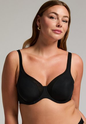 Frau trägt einen schwarzen Bügel-BH mit verstellbaren Trägern und posiert vor einem einfarbigen hellgrauen Hintergrund.