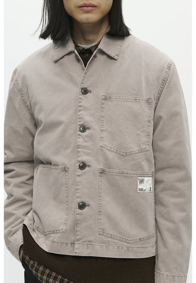 Licht taupe canvas jack met een knoopsluiting, twee opgestikken zakken en een label op de onderste zak. Gestructureerde stof met zichtbare stiksels.