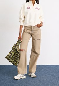 Felpa crema con zip a metà con ricamo '59', abbinata a jeans beige strappati e una borsa a tracolla con stampa leopardata verde. Sneakers bianche completano il look.