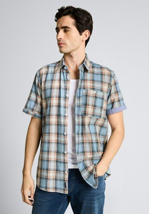 Junger Mann, der ein kurzärmliges, blau-beiges kariertes Hemd über einem weißen Tanktop trägt, mit einer Hand in der Jeans-Tasche.