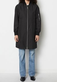 Persona con una chaqueta bomber negra de cremallera larga, vaqueros rectos de color azul claro y botas negras hasta el tobillo, de pie contra un fondo blanco.