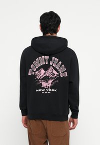 Sort hættetrøje med et stort grafisk print med bjerge og "TOMMY JEANS" i bue, tekst i pink, placeret på ryggen.