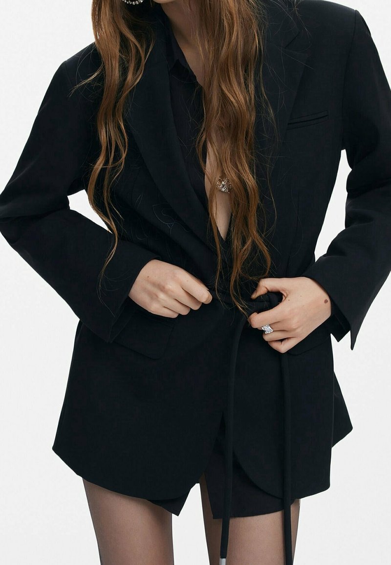 Femme aux longs cheveux portant un blazer noir oversize, attachant la ceinture, avec une bague et des boucles d'oreilles visibles.