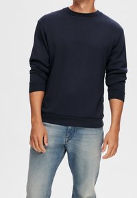 Mörkblå bomullströja med rund hals, långa ärmar och ribbade muddar och nederkant. Slät textur, avslappnad passform. Kombinerad med ljusblå jeans.