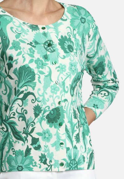 Sieviete valkā tirkīza un baltas krāsas ziedu raksta pogotu kardiganu ar apaļu kakla izgriezumu un garām piedurknēm, roka kabatā.