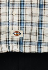 Πουκάμισο με κουμπιά σε καρό μπλε, μαύρο και καφέ χρώμα, με ορατό πλακέτα με το λογότυπο Dickies κοντά στη βάση, φορεμένο πάνω από μαύρο παντελόνι.