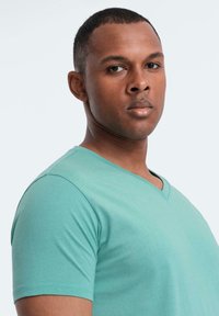 Mladý muž s krátkými vlasy, oblečený v teal V-krku tričku, hledící přímo vpřed s neutrálním výrazem na světlém pozadí.