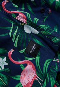 Tengerkék ing flamingó mintával, élénk rózsaszín flamingókkal, zöld pálmalevelekkel, fehér virágokkal és fekete márkacímkével. Sima szövettextúra.