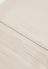 Bežinis odinis piniginė su iškilniu "Valentino" logotipu, turinti kelis kortelių skyrius ir matomus siuvinėjimo detales. Lygus paviršius.