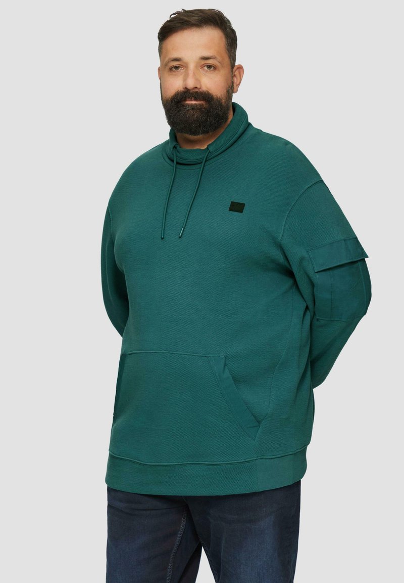 Sweat-shirt à capuche teal avec une poche kangourou et un détail de manches boutonnées sur le côté, fabriqué à partir d'un tissu doux et texturé, associé à un jean noir.