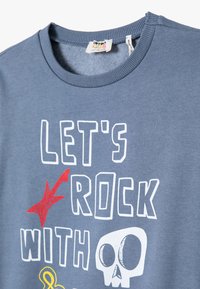 Niebieska bawełniana koszulka z tekstem "POPROSTU ROCKUJEMY Z", czerwoną gwiazdą i białą grafiką czaszki. Okrągły dekolt i krótkie rękawy.