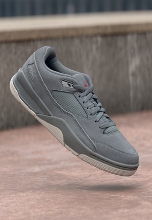 Baskets en daim gris avec lacets et panneau latéral texturé, flottant au-dessus d'une surface en béton avec un arrière-plan urbain flou.