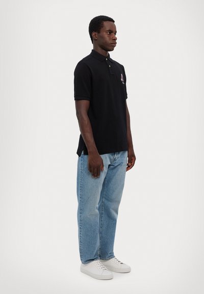Polo shirt noir à manches courtes, col boutonné et fentes latérales, associé à un jean droit bleu clair et des baskets blanches.