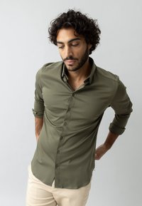 Camicia button-up verde oliva, maniche lunghe arrotolate fino ai gomiti, texture liscia, leggero luccichio, bottoni coordinati lungo il davanti.