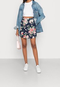 Jupe patineuse marine à fleurs roses et vertes, associée à un haut court blanc et une veste en jean. Baskets blanches et petit sac rose.