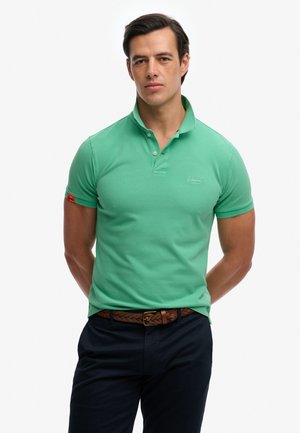 Mann trägt grünes kurzärmeliges Poloshirt, braunen geflochtenen Gürtel und dunkle Hose, steht mit Händen hinter dem Rücken.