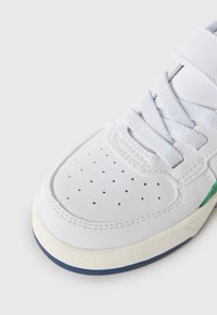 Baskets blanches avec une tige en cuir lisse, bout perforé, lacets gris et accent vert sur le côté. Elles présentent une semelle en caoutchouc texturée.