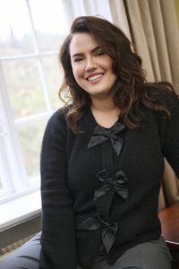 Naeratus näoga naine, kellel on pikad pruunid juuksed, istub akna ääres, kandes musta kardigani, millel on kolm suurt musta lipsu, ja halli pükse.