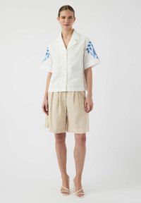 Hvit kortermet skjorte med blå bladbroderi på skuldrene, sammen med beige plisserte shorts. Modell iført hvite stroppete hæler.