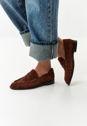 Persoon met opgerolde blauwe jeans en bruine suède loafers, staand op een glad wit oppervlak met één voet lichtjes opgetild.